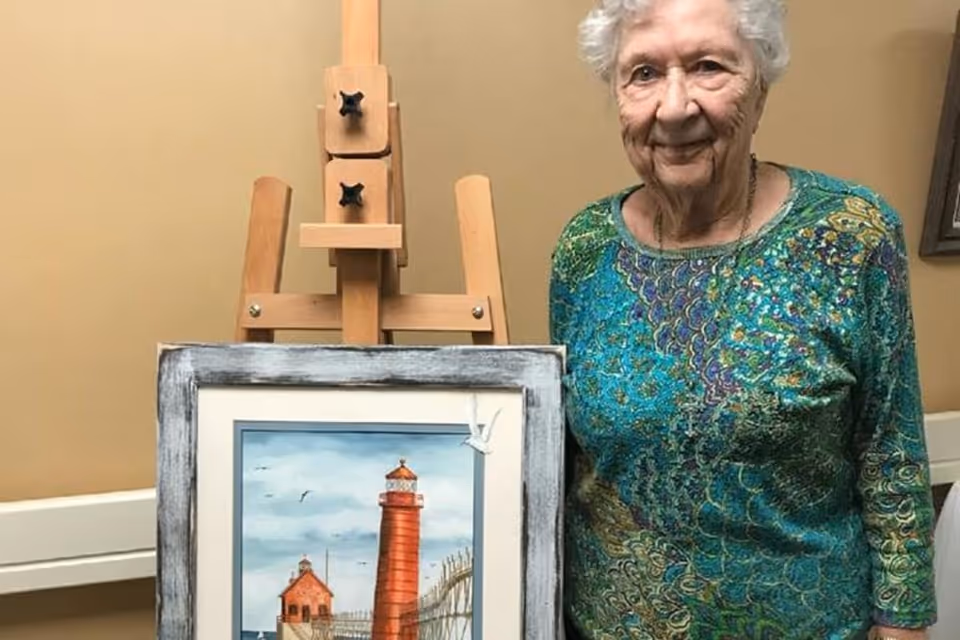 An elderly woman with short white hair wearing a green patterned top stands next to an easel holding a framed painting of a red lighthouse and buildings by the water under a cloudy sky.