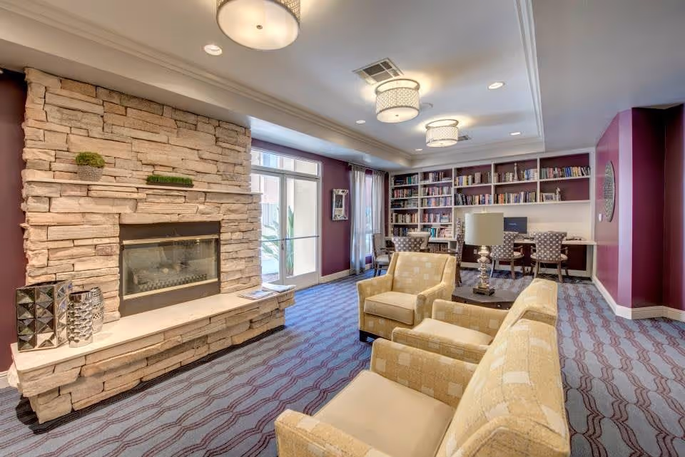 A cozy living room area with a stone fireplace on the left wall, decorated with small plants and vases. The room has purple walls and a patterned carpet. There are three beige armchairs arranged around a small table with a lamp. In the background, there is a built-in bookshelf filled with books and a desk with three chairs and a computer. Large windows with curtains allow natural light to enter the room.