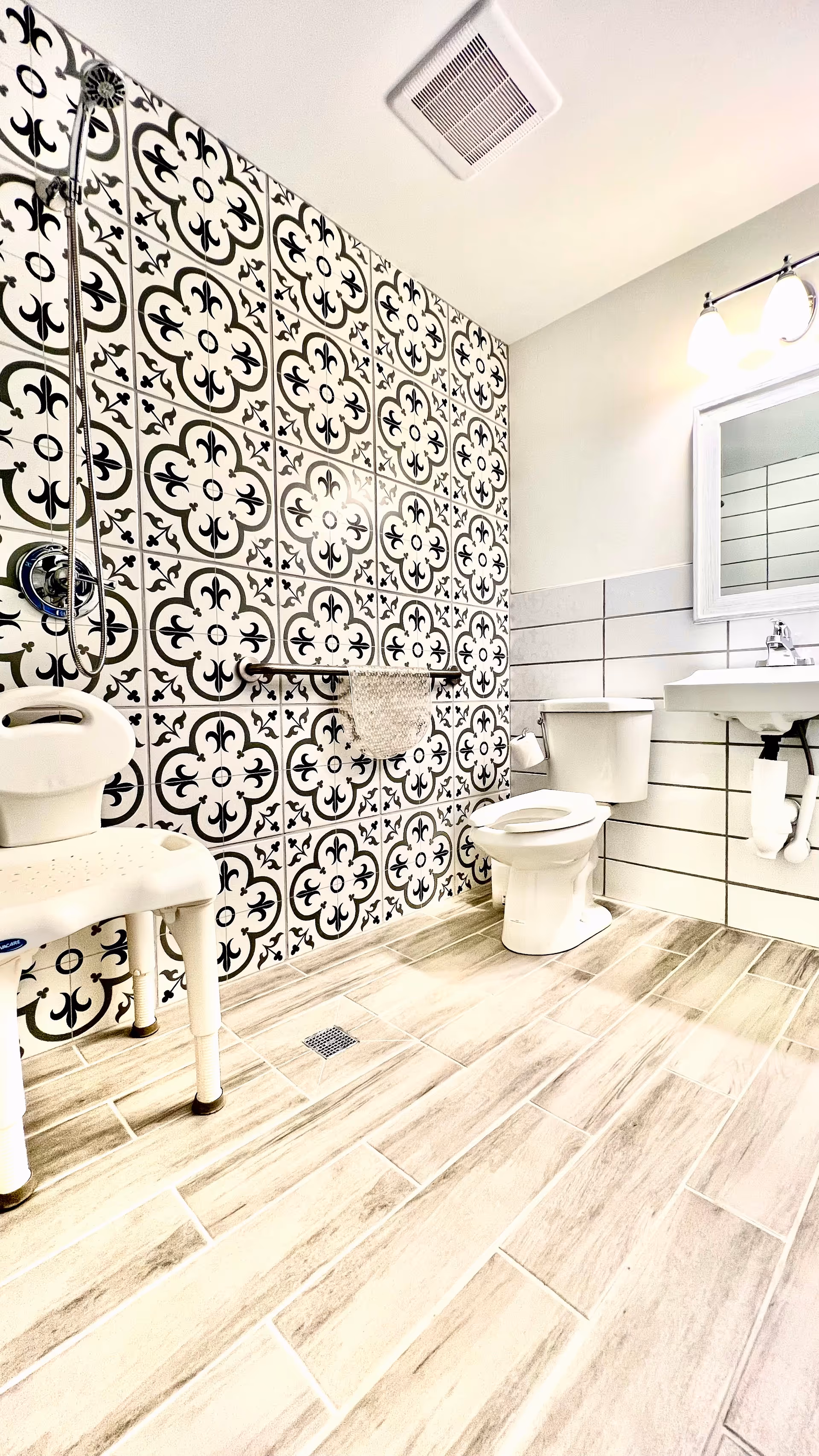 A bright and clean senior living bathroom featuring a patterned black and white tiled wall with a showerhead and grab bar. There is a white shower chair, a white toilet, a white sink with a mirror above it, and light wood-look floor tiles.