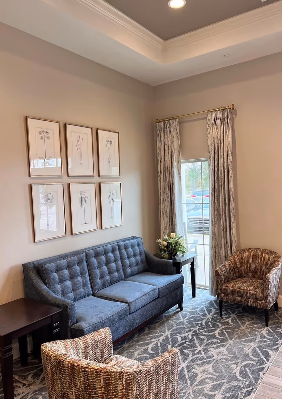 A cozy living room area with a blue tufted sofa, two patterned armchairs, a side table with a flower arrangement, six framed botanical prints on the wall, and a window with patterned curtains letting in natural light.