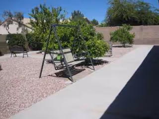 Outdoor courtyard with a metal swing bench, gravel ground, trees, and a concrete walkway.