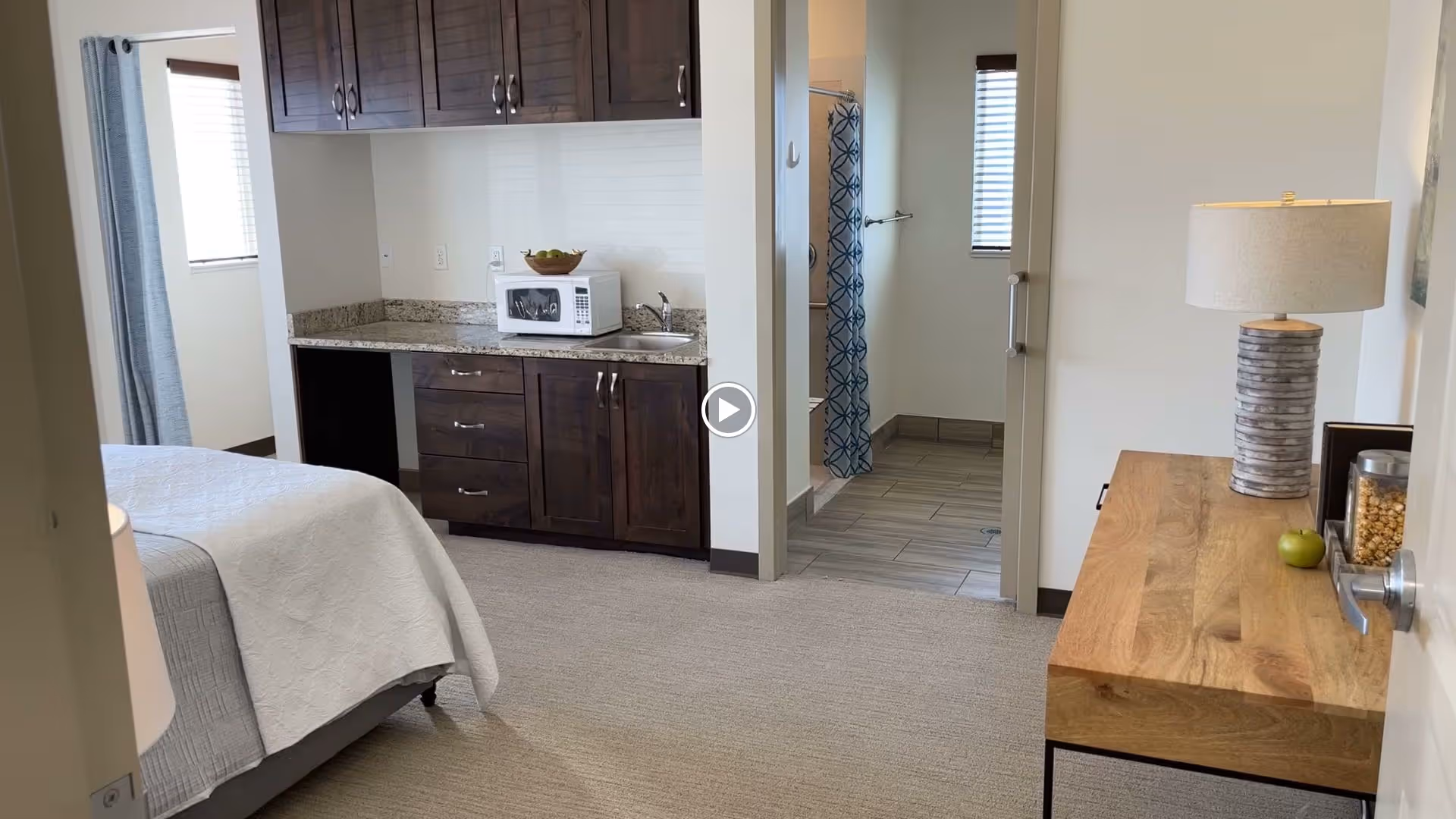 A bedroom area in an assisted living facility featuring a bed with white bedding, a wooden cabinet with a microwave and sink, and a wooden dresser with a lamp and some snacks. A bathroom with a shower and blue patterned curtain is visible through an open doorway.