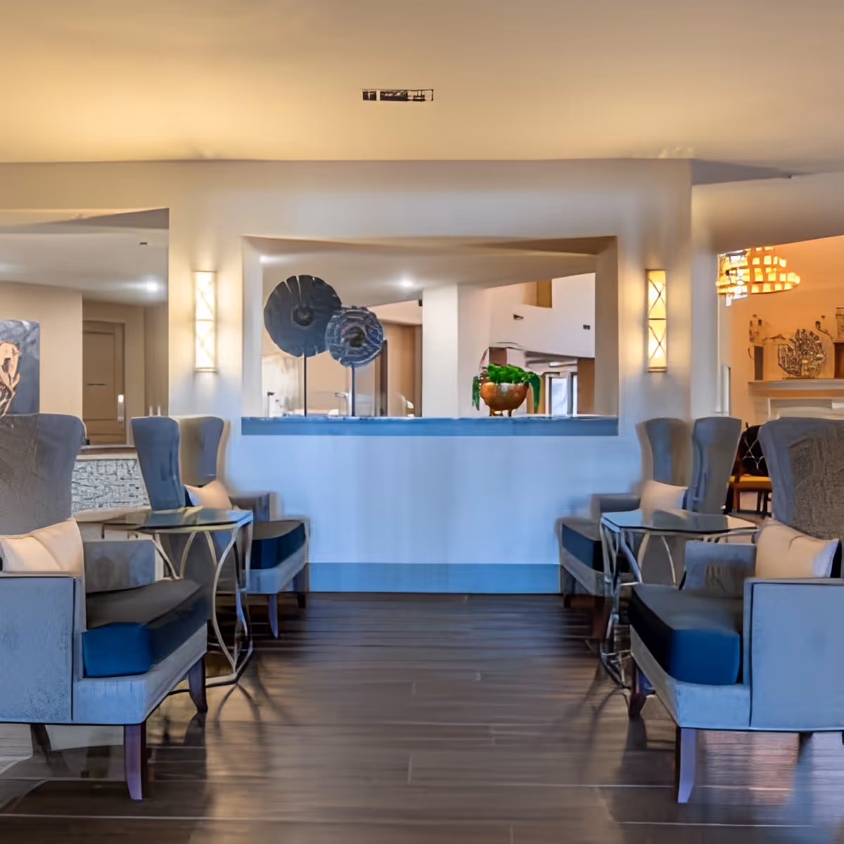 A modern and elegant lounge area with four blue upholstered armchairs arranged in pairs facing each other, separated by small glass-top side tables. The room features warm lighting from wall sconces and a chandelier in the background, with decorative art pieces and a potted plant on a ledge dividing the space.