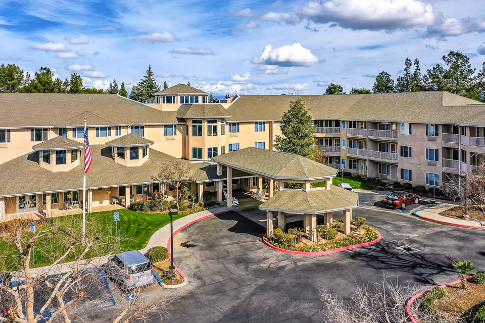 Exterior view of Solstice Senior Living at Bakersfield, showing a large multi-story building with beige walls and a brown roof. The entrance features a covered drop-off area with a hexagonal roof, surrounded by landscaped greenery and a circular driveway. Several cars are parked nearby, and an American flag is visible on a flagpole in front of the building. Trees and a partly cloudy sky are in the background.