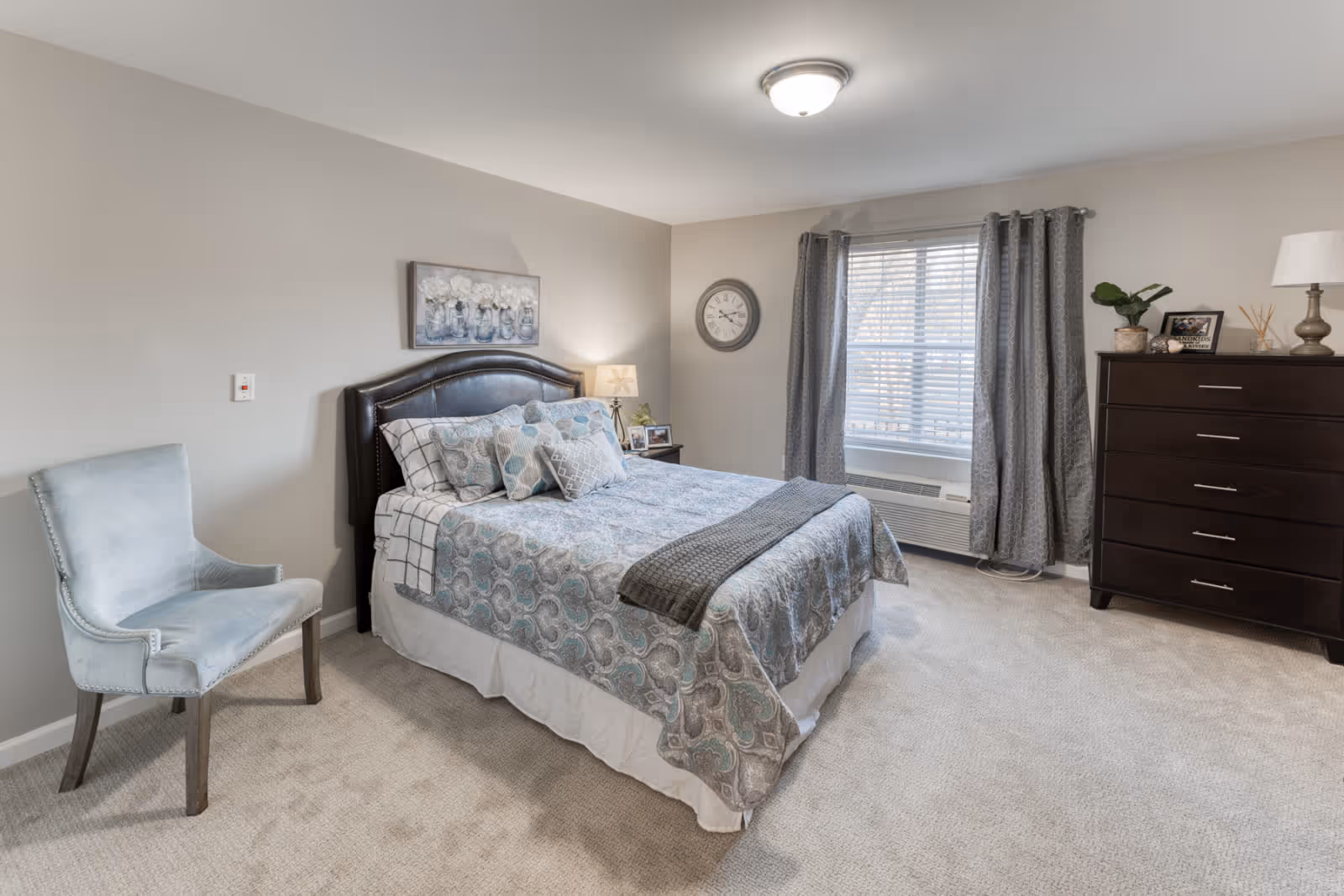 Well-lit furnished bedroom with a bed, upholstered chair, dresser, window with curtains, and neutral decor.