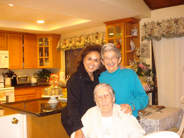 Three women smiling and posing together in a warmly lit kitchen with wooden cabinets, a granite countertop island, and floral curtains. One elderly woman is seated while two others stand behind her, all appearing happy and close.