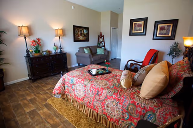 Furnished bedroom with a patterned red bedspread, wooden dresser and lamps, and a small seating area with a loveseat and chair.
