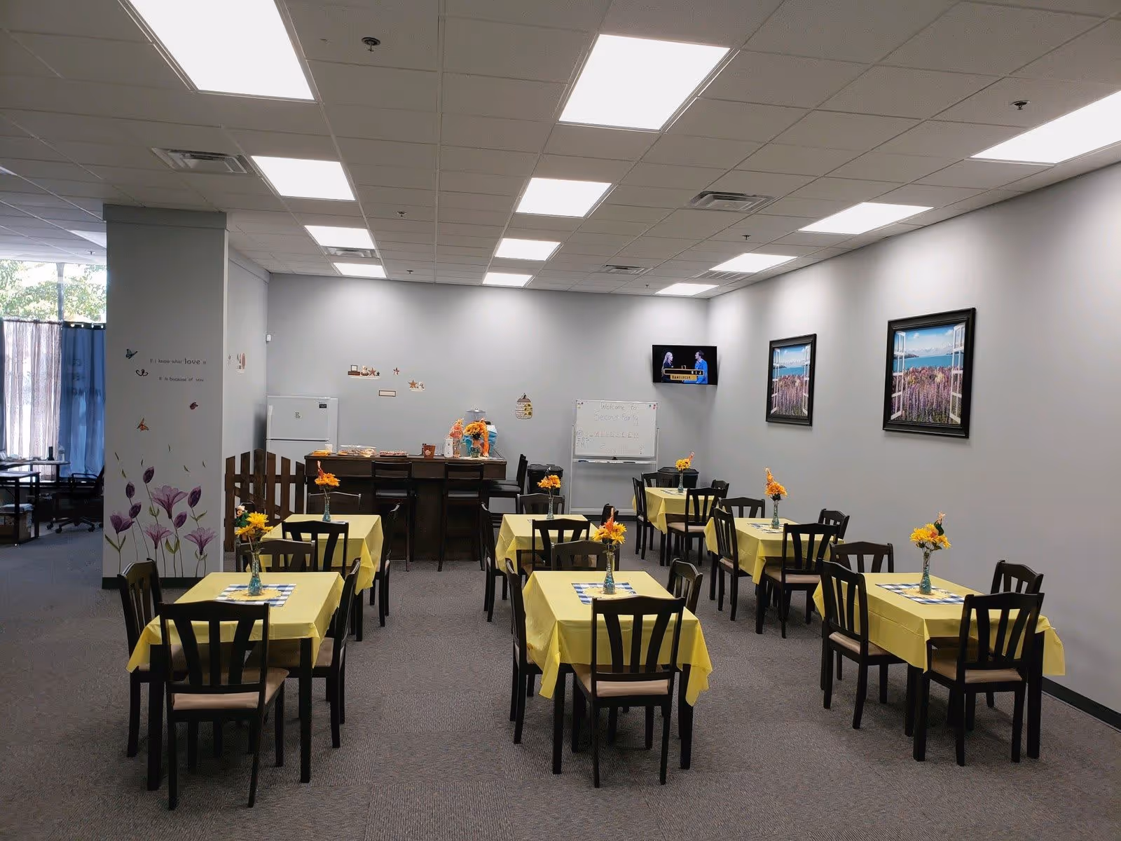 A dining room with several tables covered in yellow tablecloths, each with a small vase of yellow flowers. There are dark wooden chairs around the tables. The room has gray walls with two framed pictures and a small TV mounted on one wall. A whiteboard and a counter with a refrigerator and some items are visible at the back. The ceiling has bright fluorescent lights.