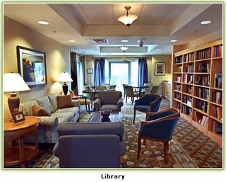 A cozy senior living facility library with comfortable seating including sofas and armchairs arranged around a coffee table. There are bookshelves filled with books along one wall, a carpeted floor with a patterned rug, and a window with blue curtains letting in natural light. The room is warmly lit with table lamps and ceiling lights.