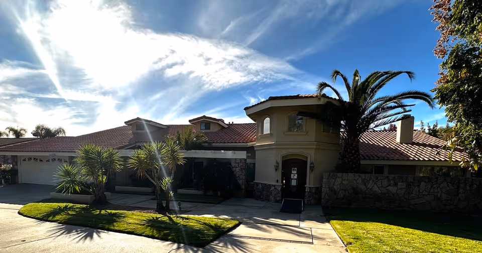 Exterior view of a single-story building with a red tile roof, stone accents, and a front entrance surrounded by palm trees and green lawn under a partly cloudy sky.