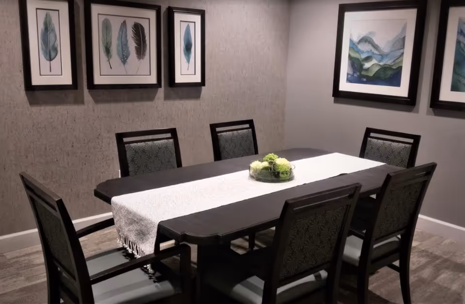 A dining room with a dark wooden rectangular table covered by a white table runner and a centerpiece of green and white flowers. Six chairs with patterned upholstery surround the table. The walls are decorated with framed artwork featuring feathers and abstract landscapes.