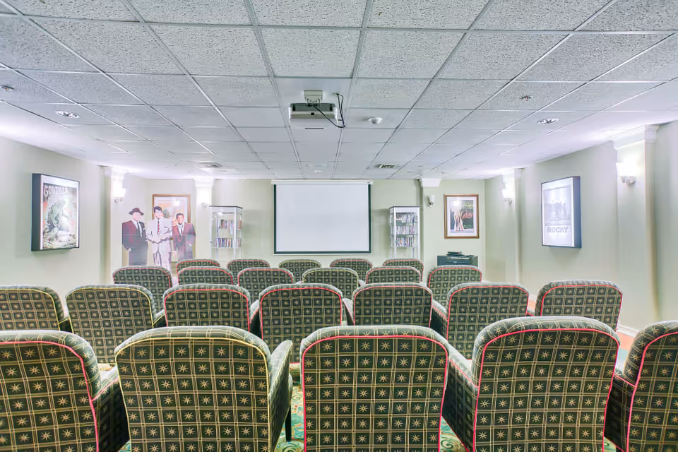 A small movie theater room with multiple rows of green patterned armchairs facing a white projection screen. The walls are light-colored and decorated with framed movie posters including Godzilla and Rocky. There are two glass cabinets with DVDs or books on either side of the screen.