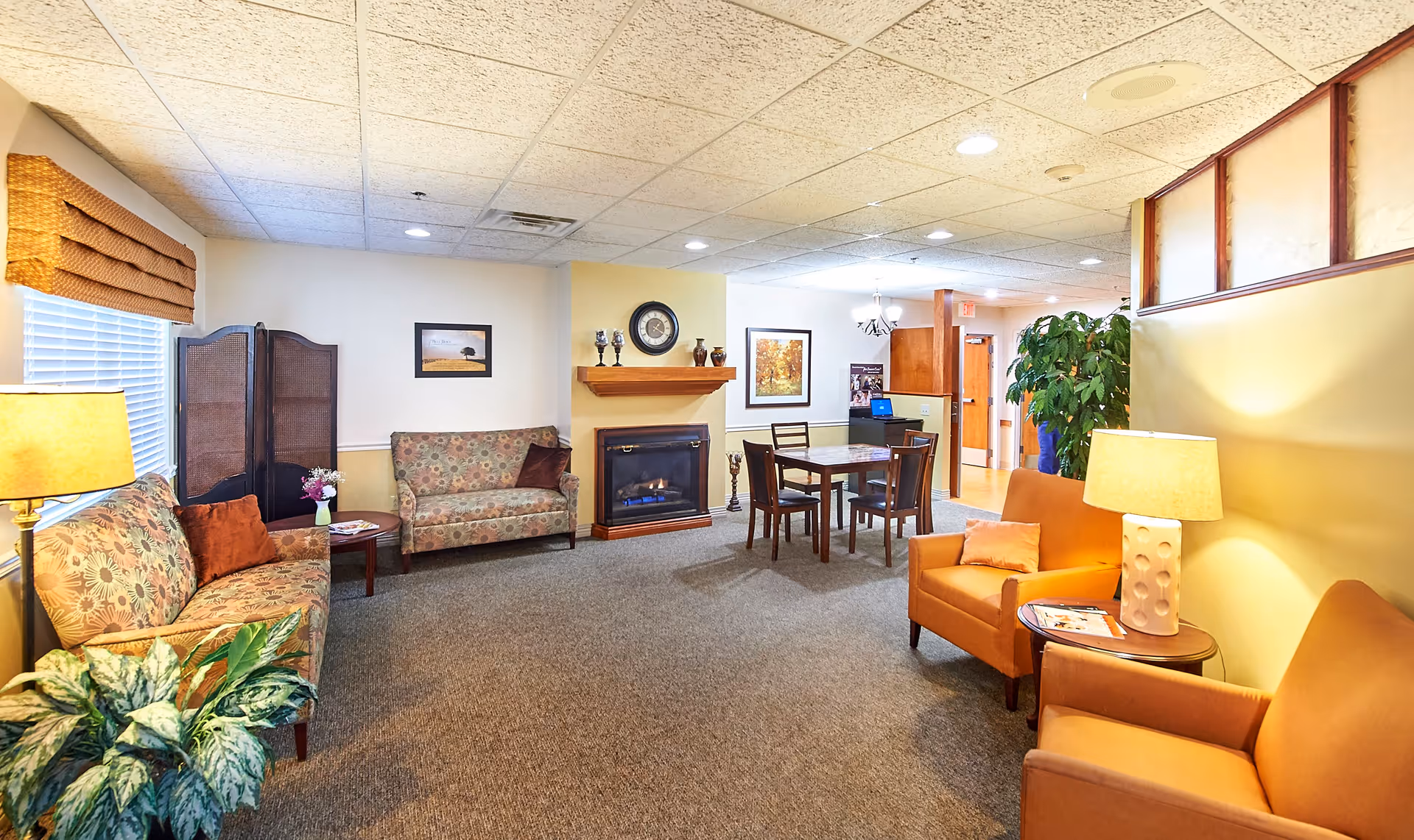 A cozy senior living facility common area with patterned sofas, orange armchairs, a small dining table with four chairs, a fireplace with decorative items on the mantle, framed artwork on the walls, and soft lighting from table lamps. There is a plant near the entrance and a folding screen by the window.