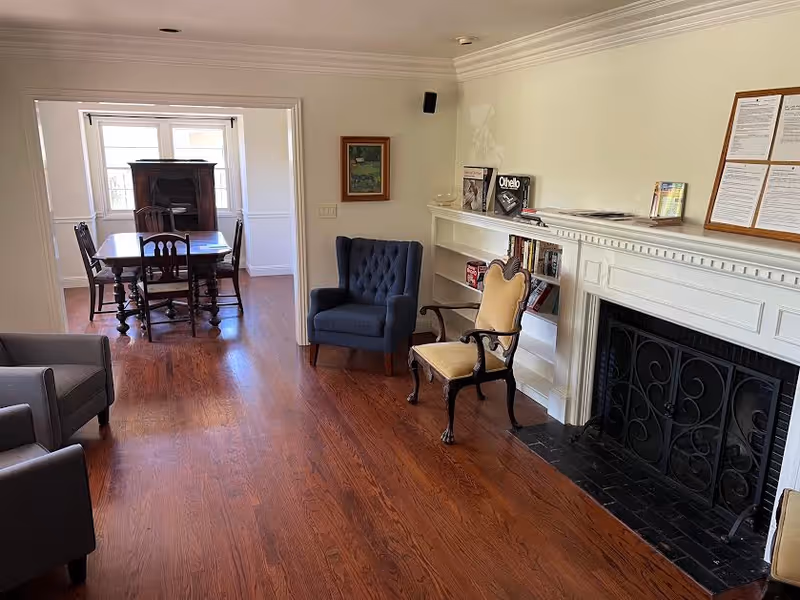 A bright living room with hardwood floors, a fireplace and bookshelves, several chairs, and a dining table visible through an open doorway.