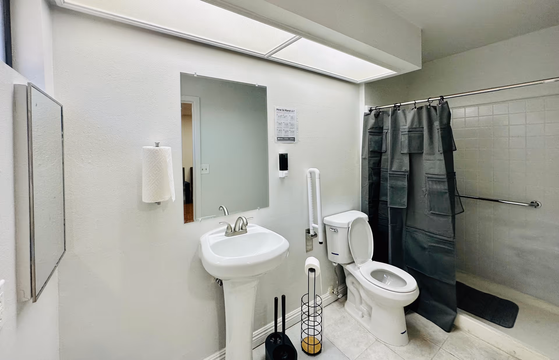A clean bathroom with a white pedestal sink, a mirror above it, a toilet with the lid open, a black shower curtain with pockets, a towel holder with a paper towel roll, and a toilet brush and plunger set on the floor. The walls are white and the floor is tiled.