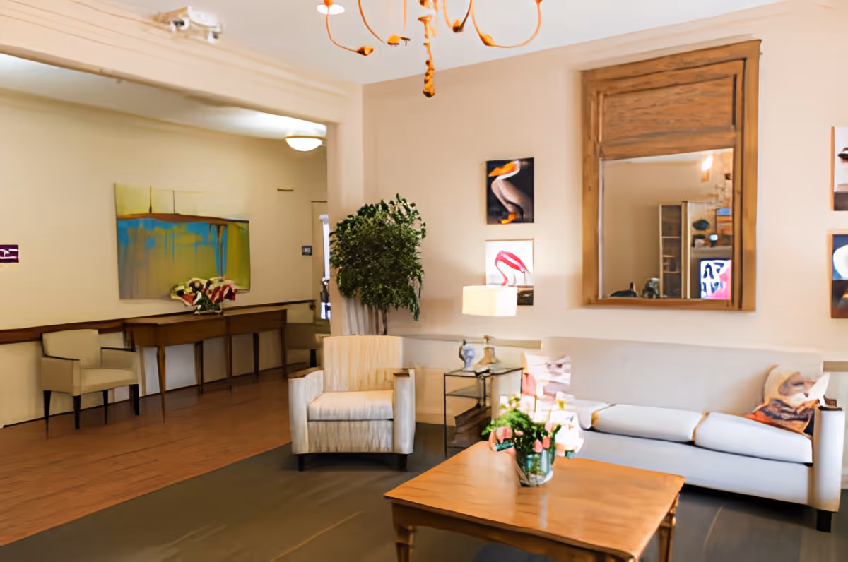 A cozy living room area with a beige armchair, a white sofa with decorative pillows, a wooden coffee table with a vase of flowers, a large wooden-framed mirror on the wall, and several pieces of artwork. There is also a side table with a lamp and a potted plant in the corner.