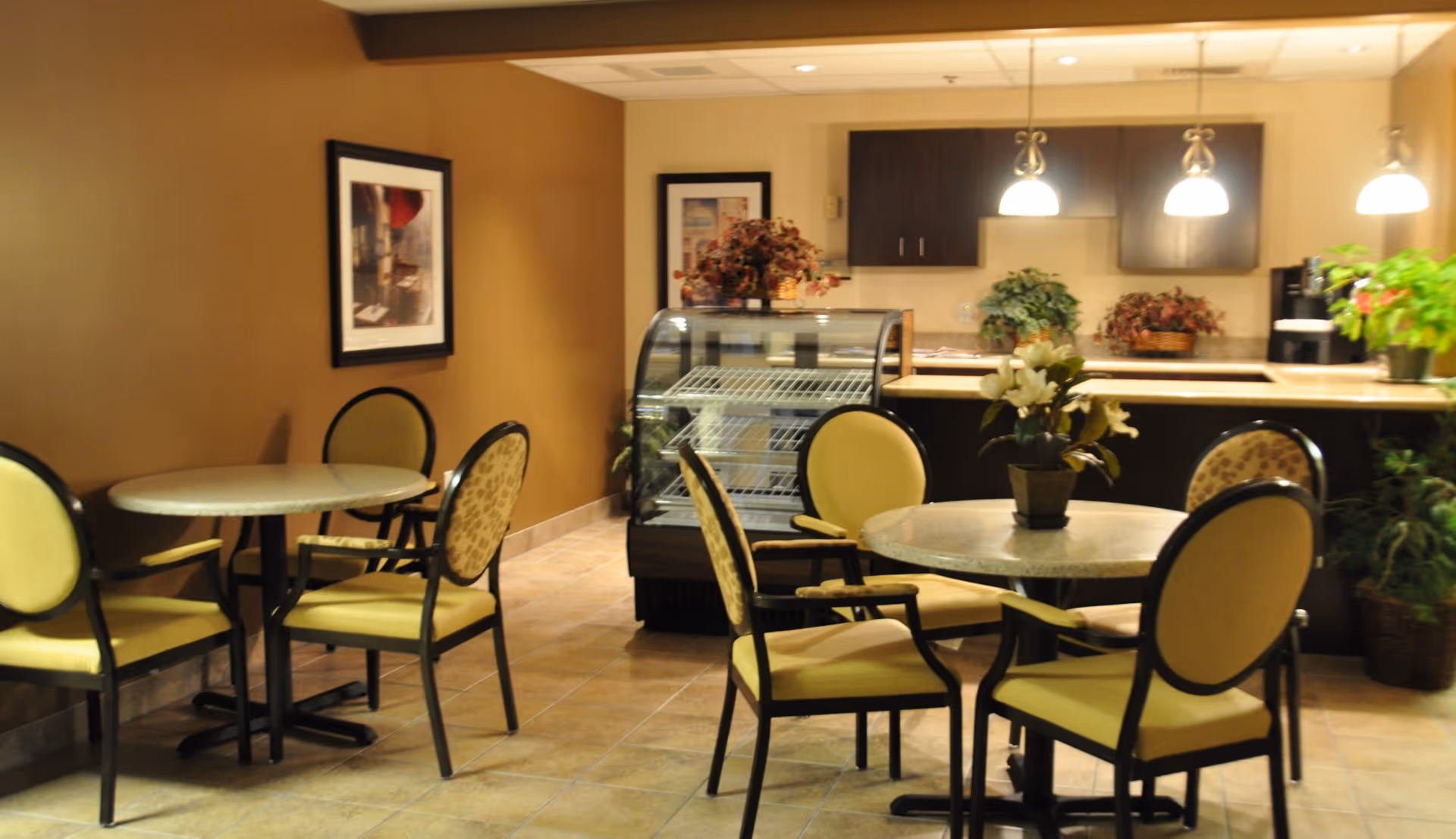 Dining area with round tables and yellow-upholstered chairs in front of a service counter and glass display case.