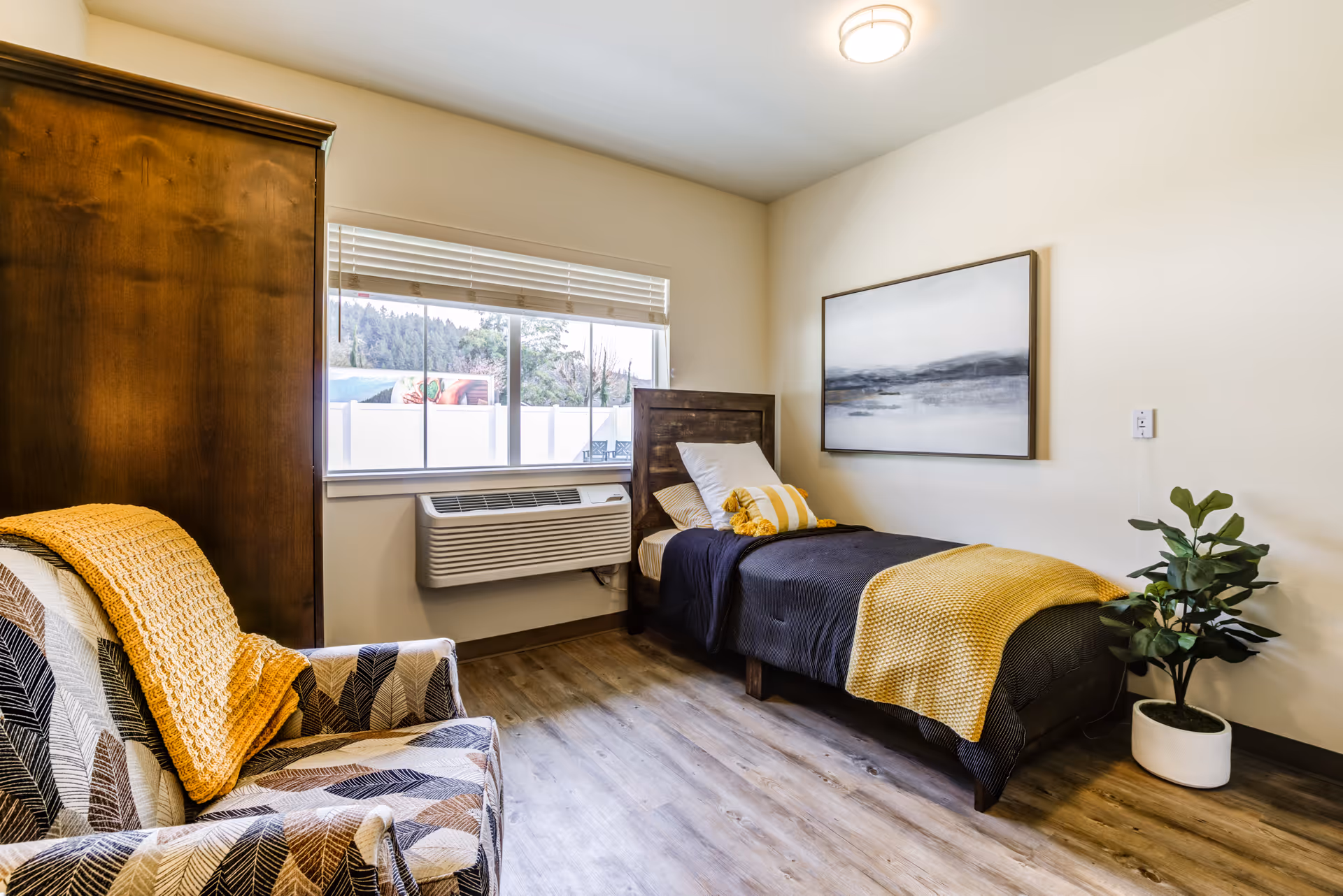 A cozy bedroom with a single bed dressed in dark bedding and a yellow throw blanket. There is a patterned armchair with a yellow knitted throw draped over it. A large wooden wardrobe stands next to a window with blinds, and a potted plant is placed near the bed. A framed landscape painting hangs on the wall above the bed. The room has wood flooring and a ceiling light fixture.