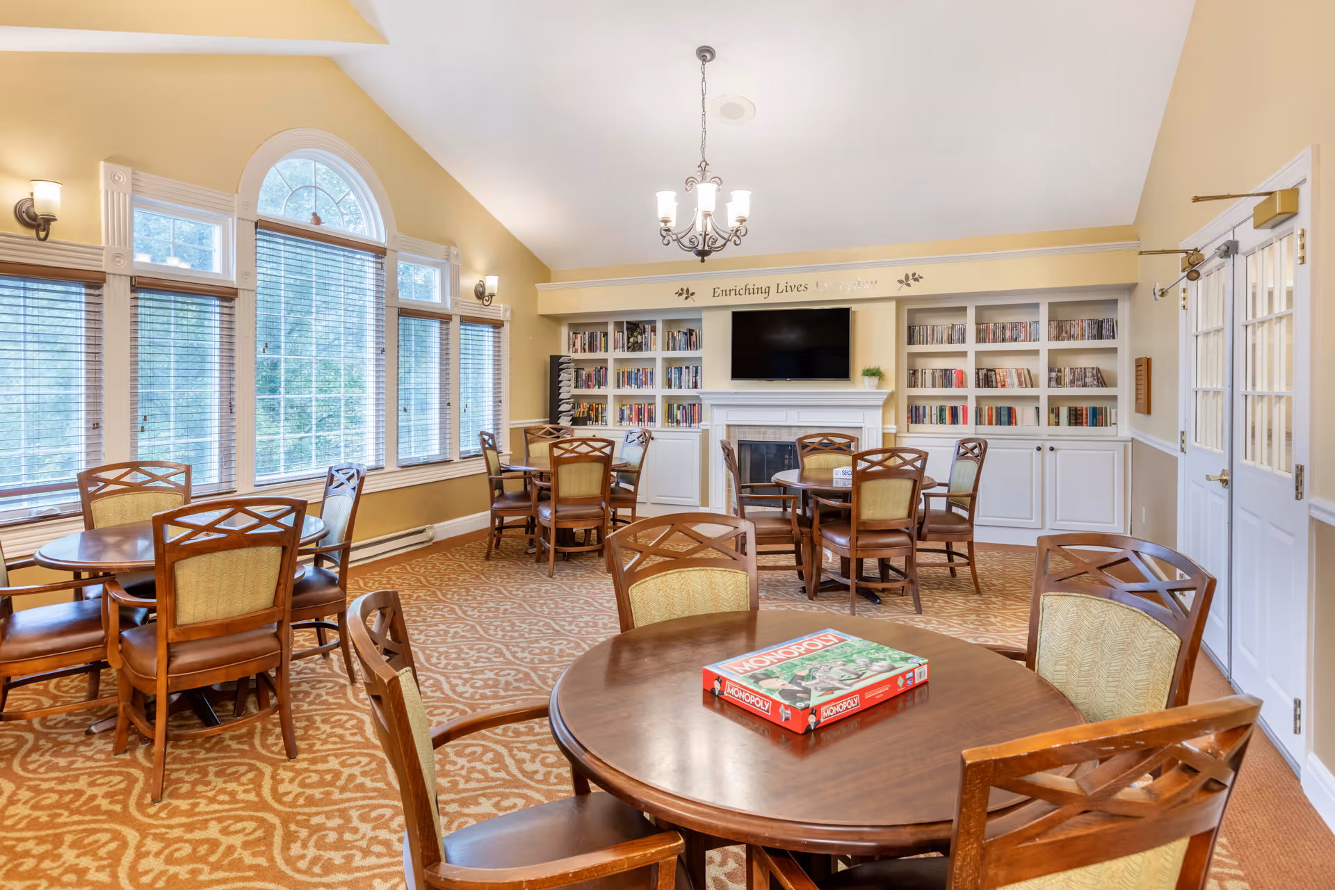 A bright and cozy common room with multiple round wooden tables and chairs arranged on a patterned carpet. Large windows with blinds allow natural light to fill the room. A built-in white bookshelf filled with books and a flat-screen TV mounted above a fireplace are visible on the far wall. A Monopoly board game box is placed on the nearest table. The walls are painted a warm yellow, and a chandelier hangs from the ceiling.