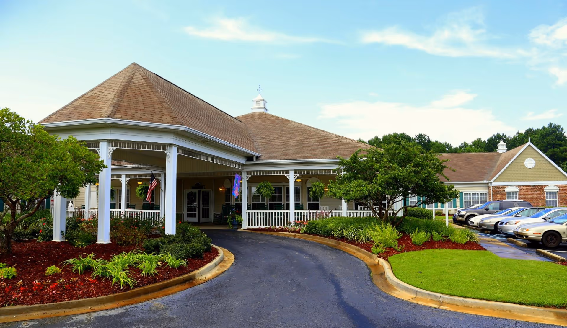 Front entrance of a senior living facility with a covered porte-cochere, landscaped beds, and parked cars along the driveway.