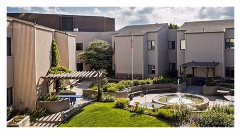 Outdoor courtyard area of a senior living facility with a circular water fountain in the center, surrounded by greenery, benches, and a pergola with seating. The building exterior is visible in the background under a partly cloudy sky.