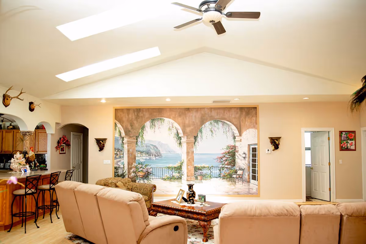 A bright living room with beige sofas facing a large wall mural depicting an ocean view with arches and greenery. The room has a ceiling fan, skylights, and an open kitchen area with bar stools on the left side. There are decorative items on the coffee table and walls, and a doorway leading to another room on the right.