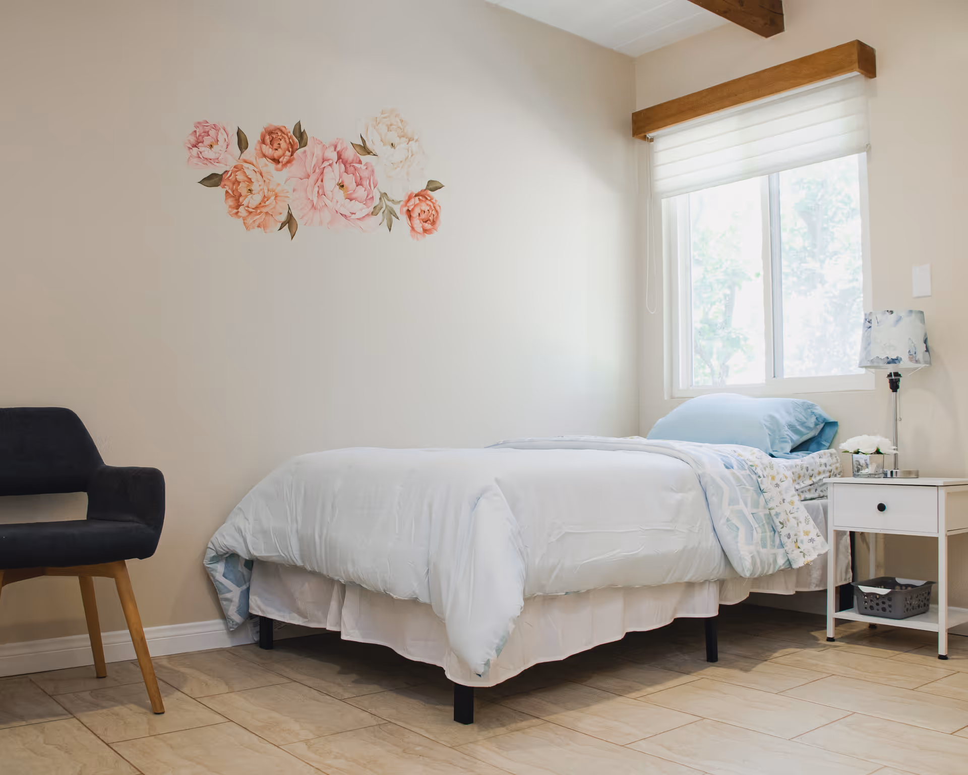 Bright single bedroom with a bed, nightstand and chair under a window and a floral wall decal.