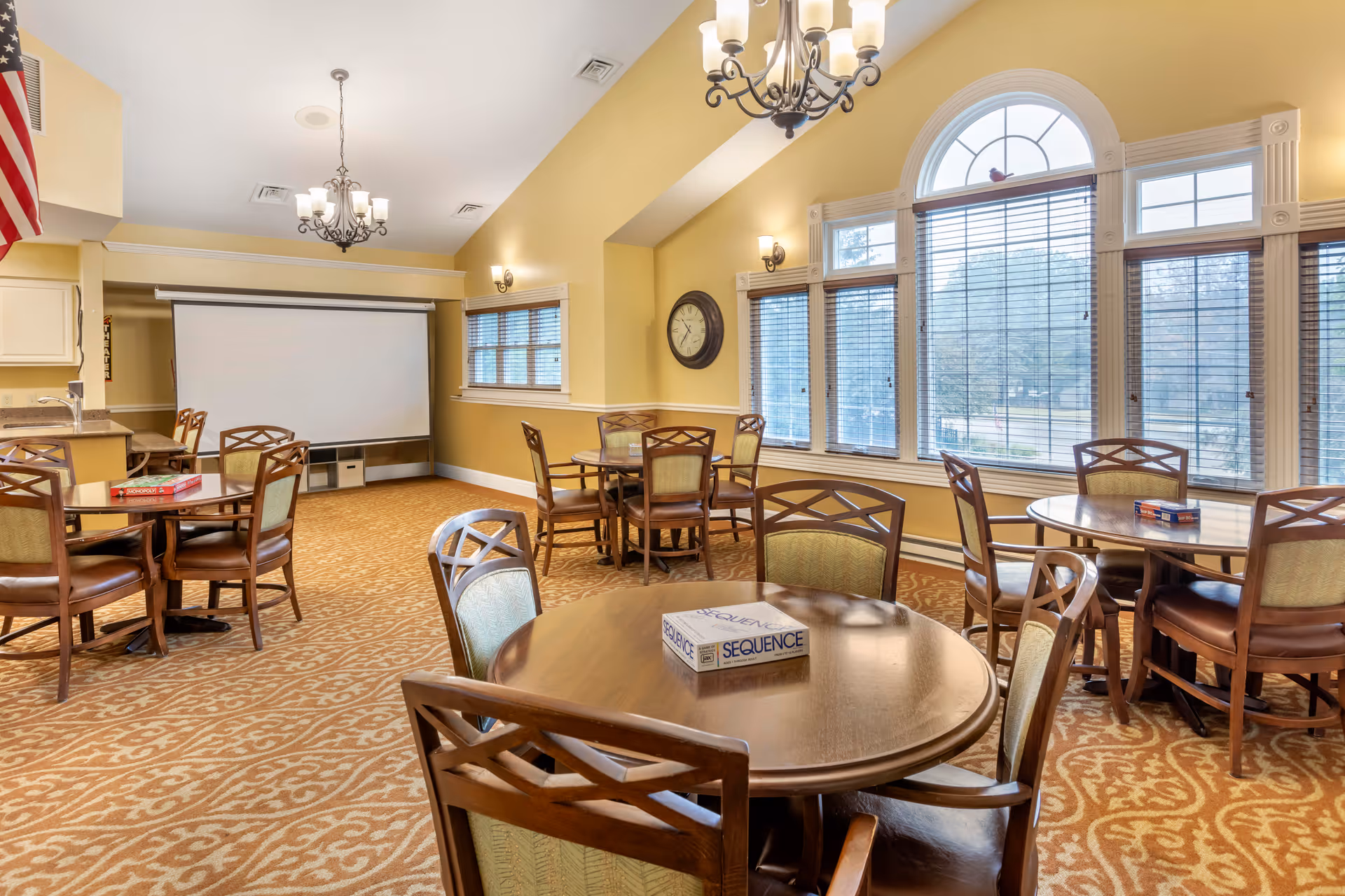A well-lit common room with multiple round wooden tables and chairs arranged on a patterned carpet. Large windows with blinds allow natural light to fill the room. A clock is mounted on the wall, and a projector screen is visible in the background. Board games are placed on some tables.