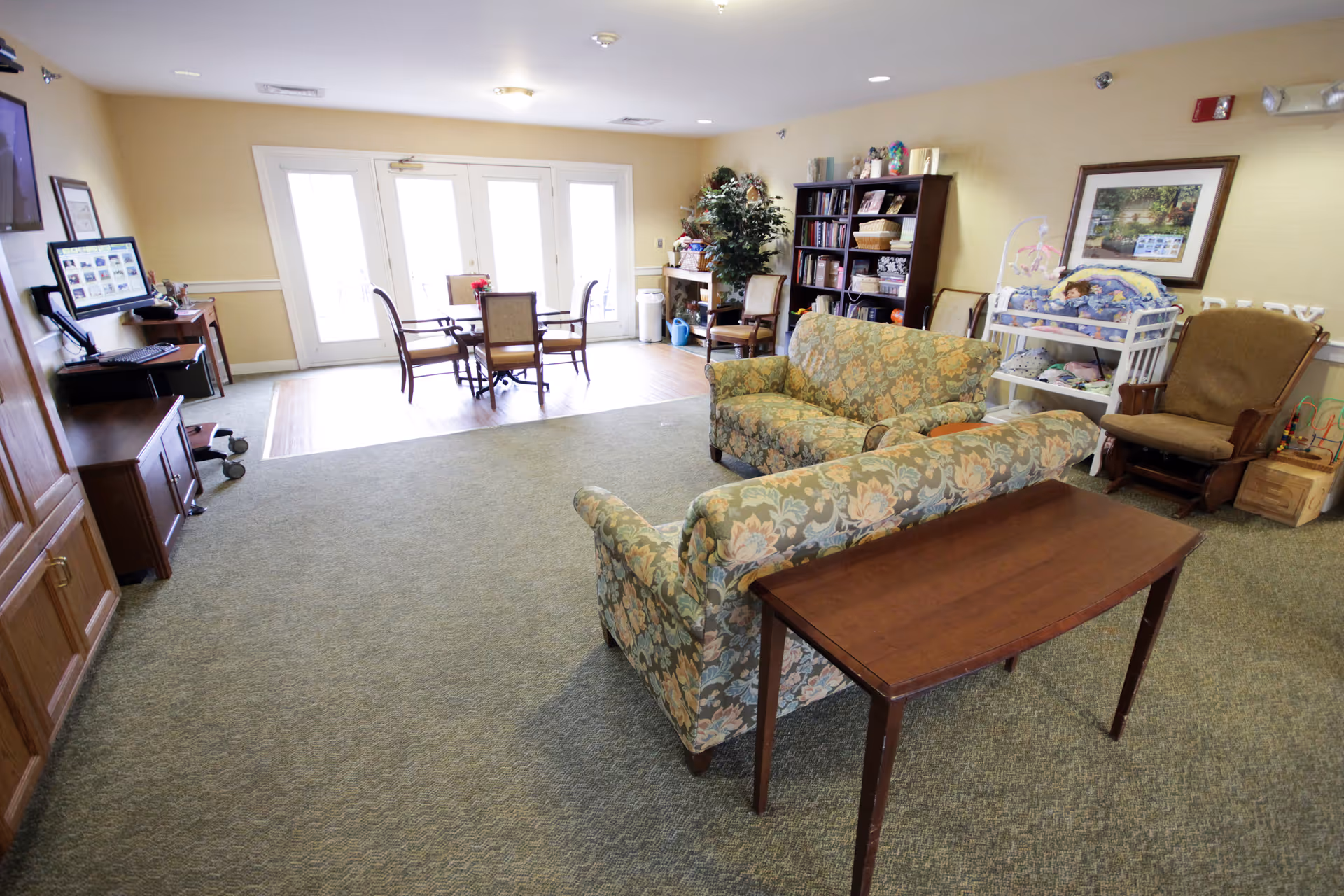A bright senior living common room with floral sofas, a wooden console table, a small dining table by glass doors, bookshelf and chairs.