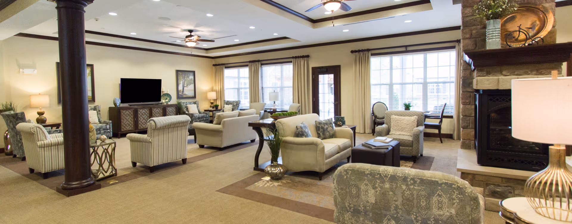 A spacious and well-lit living room area in a senior living facility with multiple upholstered armchairs and sofas arranged around a central carpet. The room features large windows with curtains, a stone fireplace with decorative items on the mantel, ceiling fans, and a flat-screen TV on a wooden cabinet. There are also side tables with lamps and decorative plants throughout the room.