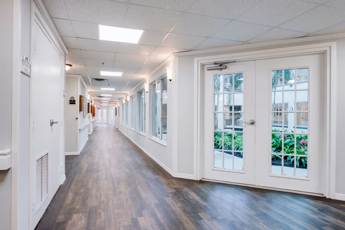 A bright, clean hallway in a senior living facility with wood-look flooring, white walls, and ceiling lights. On the right side, there are large windows and glass double doors showing a view of a garden with green plants and palm trees outside. On the left side, there are several doors and wall-mounted lights along the corridor.