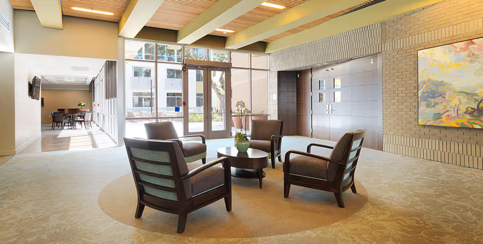 A bright and spacious common area with four cushioned armchairs arranged around a round wooden coffee table with a small plant on it. The room features large windows and glass doors letting in natural light, a beige carpet with a circular pattern under the seating area, a wooden slatted ceiling, and a colorful abstract painting on the brick wall.