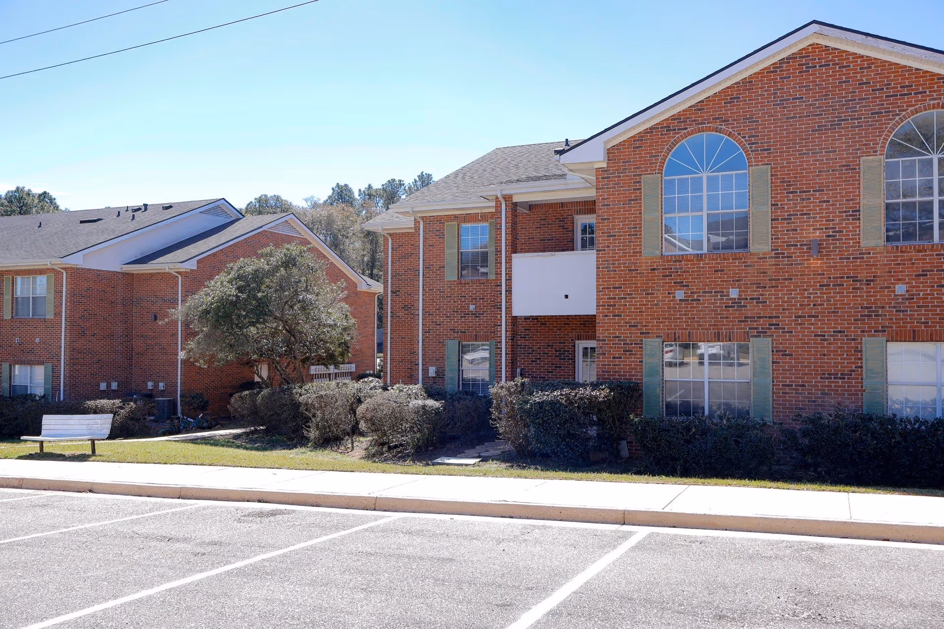 Exterior view of a senior living facility showing two connected red brick buildings with multiple windows and green shutters. There is a small balcony on the second floor of the building on the right. In front of the buildings, there are trimmed bushes, a tree, a white bench, and an empty parking lot under a clear blue sky.