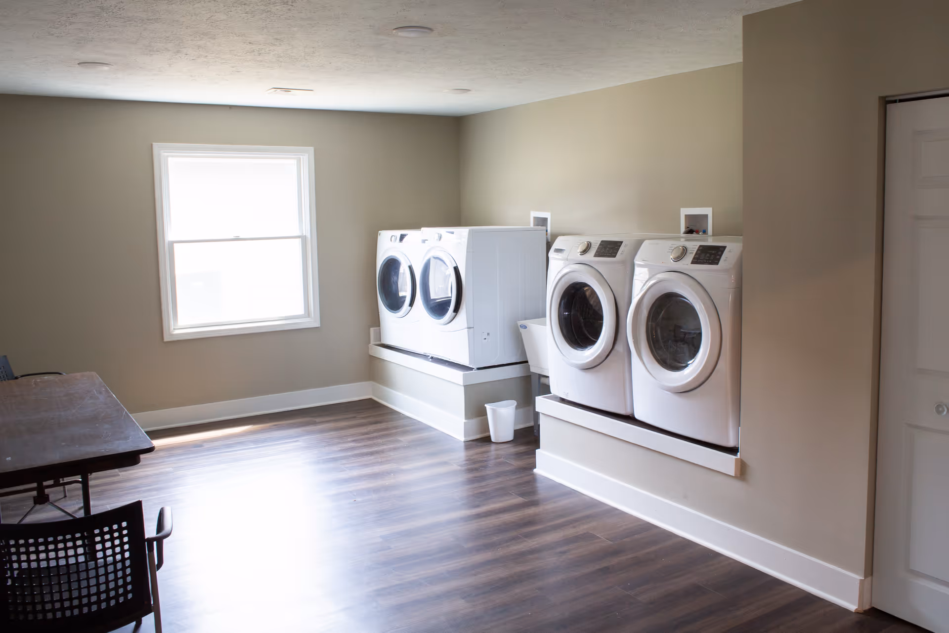 A bright laundry room with two sets of white front-loading washing machines and dryers mounted on raised platforms against a beige wall. There is a window letting in natural light, a utility sink between the machines, a small white trash bin on the floor, and a wooden table with chairs on the left side of the room. The floor is dark wood laminate.