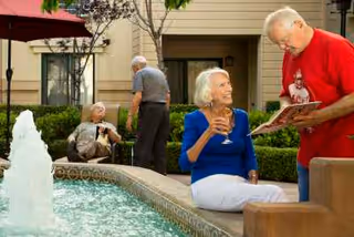 Four elderly people enjoying a sunny day in an outdoor courtyard with a water fountain. One woman in a blue top is sitting on the edge of the fountain, smiling and holding a drink, while a man in a red shirt is showing her something in a book. In the background, two other elderly men are engaged in conversation near some greenery and a building.