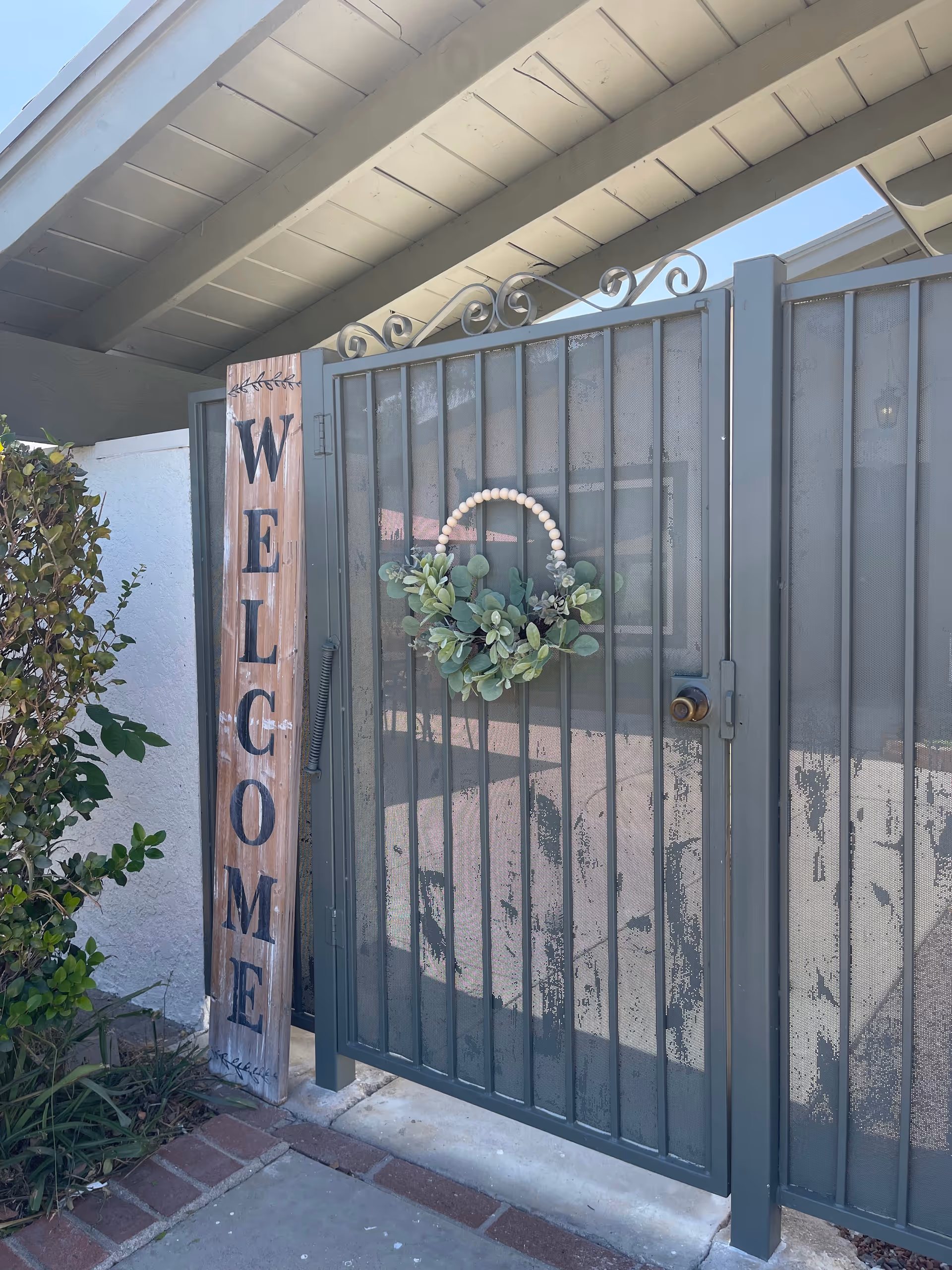 A metal front gate decorated with a circular greenery wreath and a tall vertical wooden sign reading 'WELCOME' beside it under the eaves.