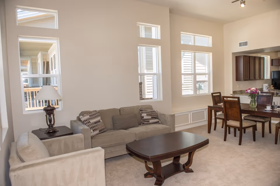 A bright living room area with beige walls and carpet, featuring a beige sofa and armchair with patterned cushions, a dark wooden coffee table, a side table with a lamp, and a dining table with four chairs. The room has multiple windows allowing natural light and an open view into a kitchen area with dark wooden cabinets.