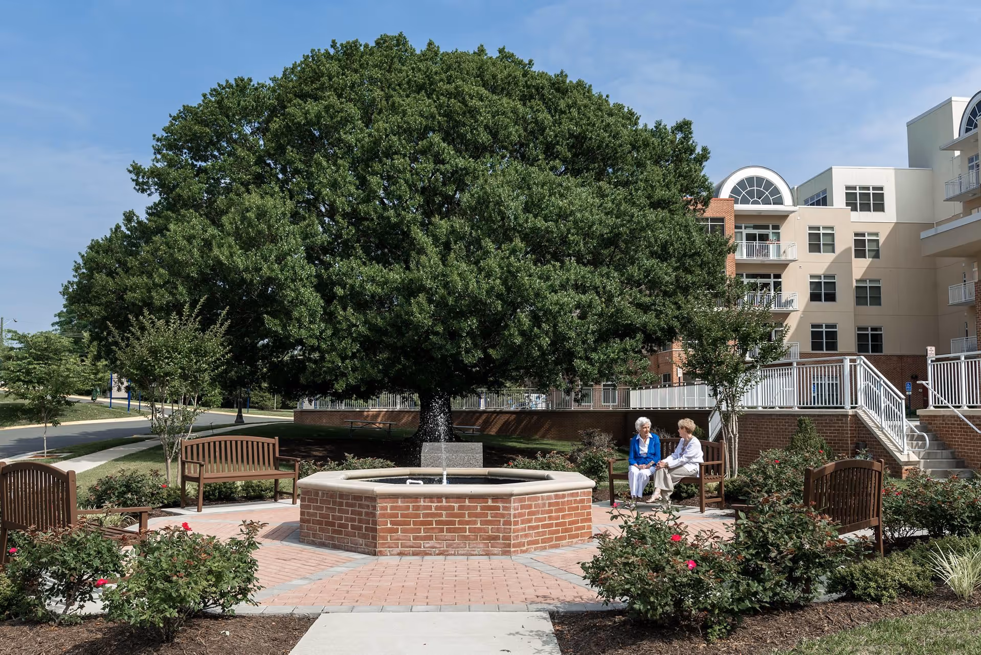 Outdoor seating area with a brick water fountain in the center, surrounded by benches and rose bushes. Two elderly women are sitting and conversing on one of the benches. Behind the seating area is a large leafy tree and a multi-story building with balconies and stairs.
