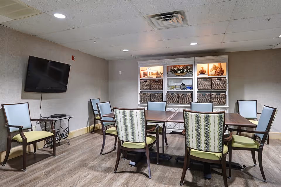 A small meeting or activity room with a rectangular wooden table surrounded by eight chairs, some with patterned backs and others with plain backs. A flat-screen TV is mounted on the wall to the left, above a small table with a device on it. A white shelving unit with decorative items and woven baskets is against the back wall. The room has wood flooring and a drop ceiling with recessed lighting.