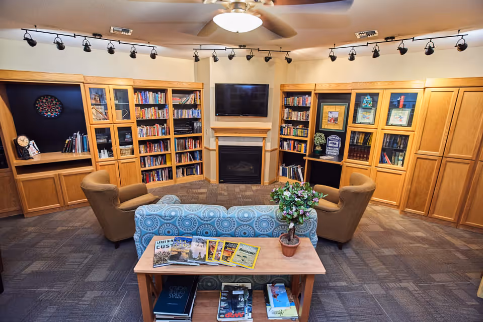A cozy library or reading room with wooden bookshelves filled with books, a flat-screen TV mounted above a fireplace, two brown armchairs, and a blue patterned sofa. A small wooden table with magazines and a potted plant is in the foreground. The room has carpeted flooring and ceiling lights.