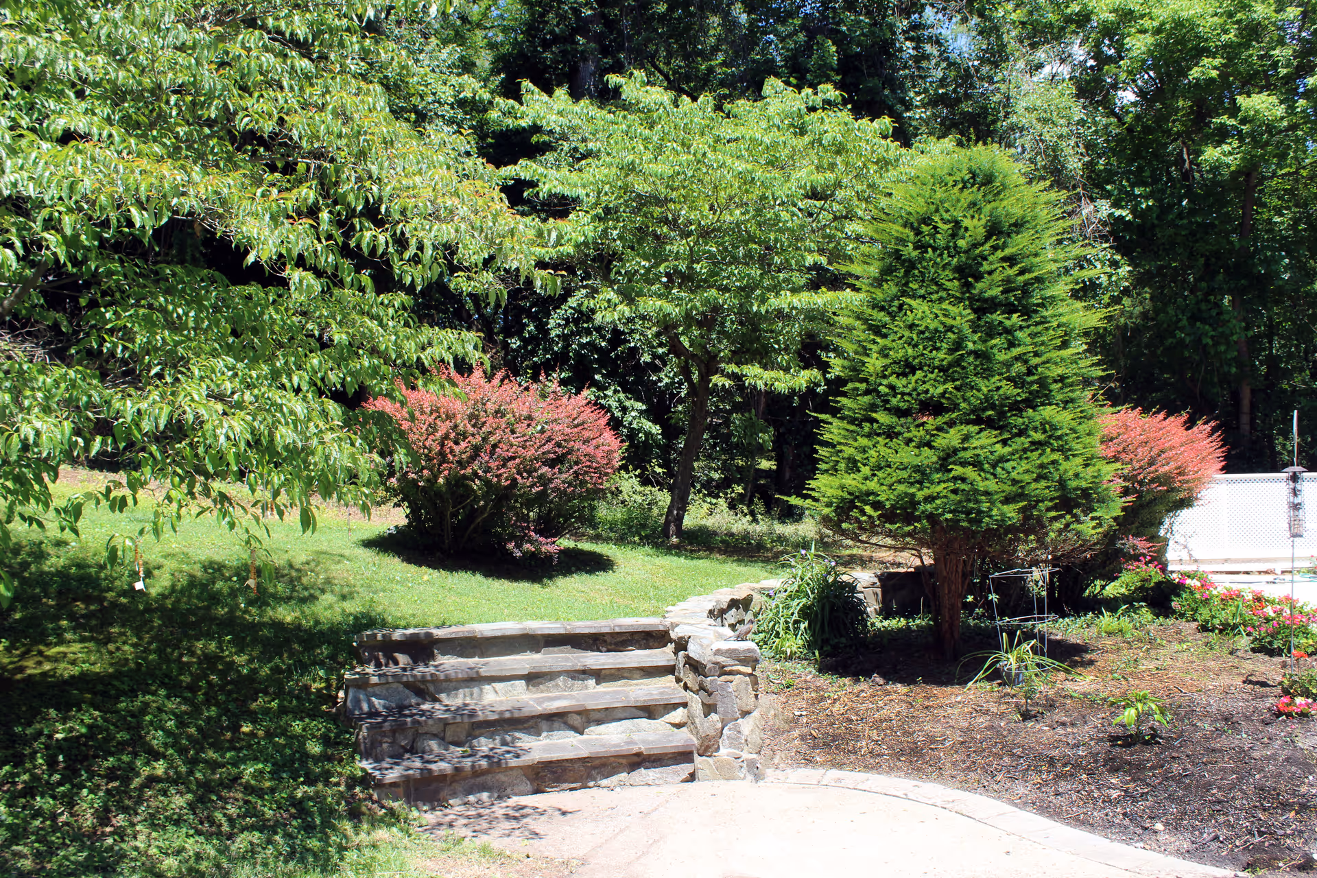Stone steps leading up to a grassy yard with trees, shrubs, and landscaped flowerbeds.