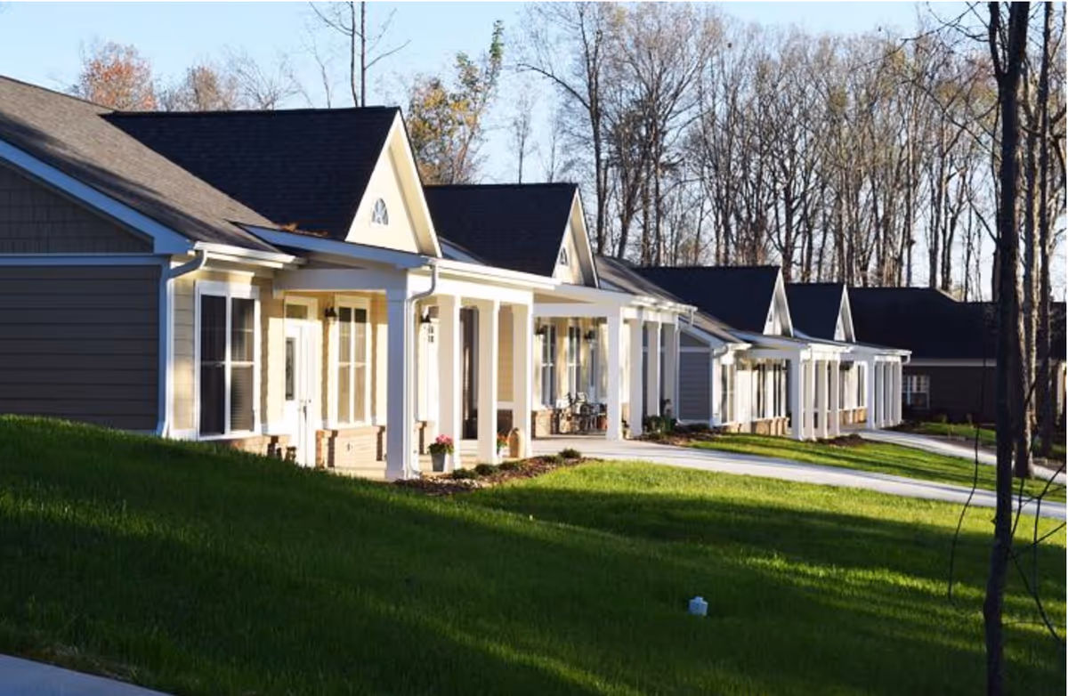 Row of single-story residential buildings with white columns and porches, surrounded by green grass and leafless trees in the background under a clear sky.