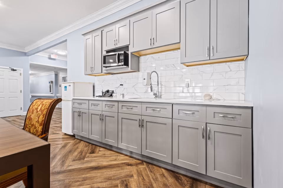 A modern kitchen area in a senior living facility featuring gray cabinets with silver handles, a white countertop, a white tile backsplash with a marble pattern, a built-in microwave, a sink with a flexible faucet, and a small white refrigerator. The floor has a wood-patterned design, and there is a wooden table with a chair that has a floral patterned cushion. The background shows a hallway with white doors and light blue walls.