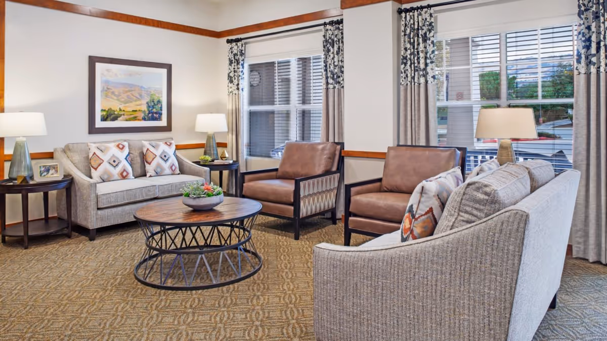 Well-lit sitting area with sofas, two leather armchairs, a round coffee table, lamps, and large windows with curtains.