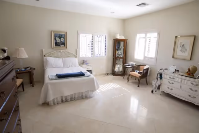 A bright and spacious bedroom with a white bed covered in white linens and a folded blue blanket on top. The room features light-colored tiled floors, two windows with white shutters, a wooden display cabinet with glass doors, a small side table with a lamp, two chairs, and a white dresser with decorative items and framed artwork on the walls.