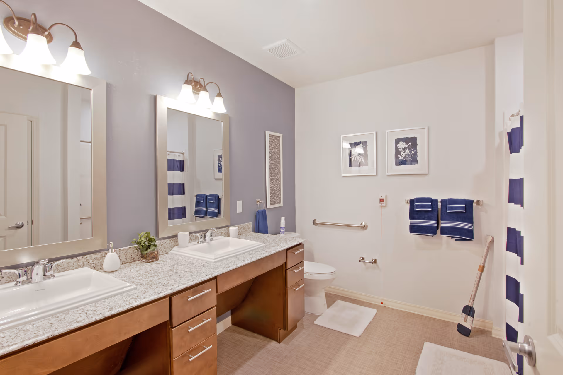 A clean and spacious bathroom with two sinks set in a granite countertop, wooden cabinets underneath, two large mirrors with light fixtures above them, a toilet with grab bars, two framed pictures on the wall, blue and white striped shower curtain, and blue towels hanging on a towel rack.