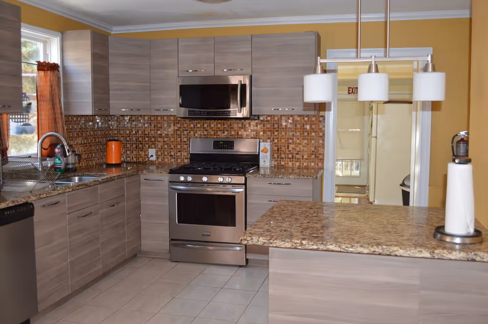 Modern kitchen with light wood cabinets, a stainless steel stove and microwave, a granite countertop island with a paper towel holder, a tiled backsplash in brown tones, and a window with orange curtains.
