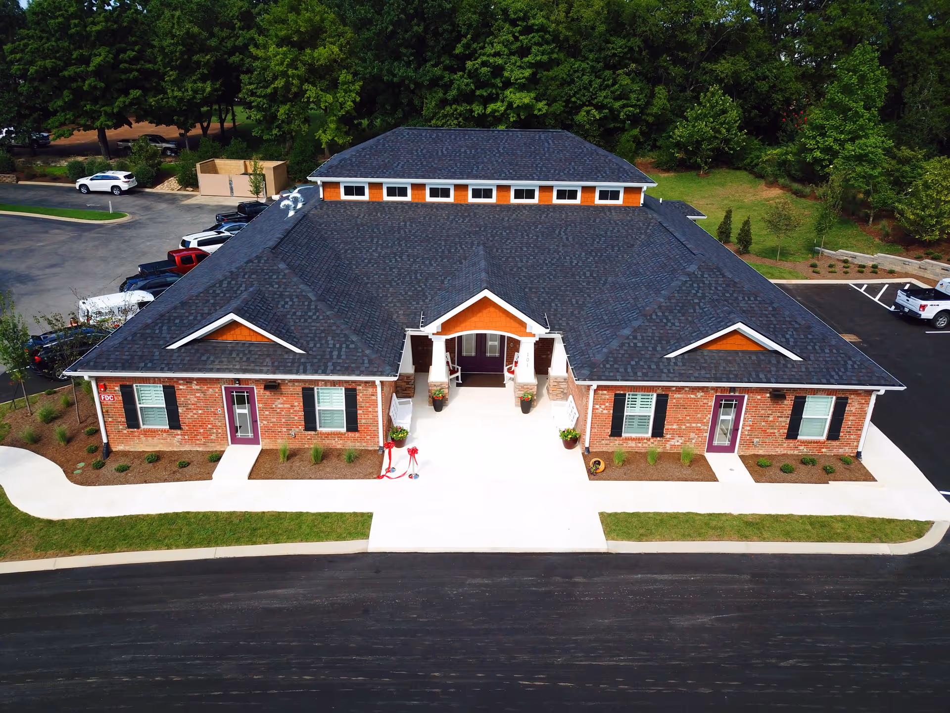 Aerial front view of a single-story brick memory care building with a central entrance, dark roof, parking lot and surrounding trees.