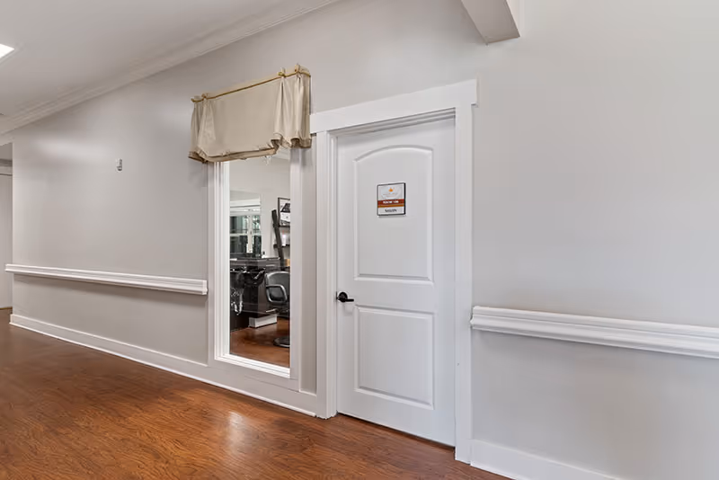 A hallway with wooden flooring and light gray walls featuring white trim and a chair rail. There is a white door with a sign labeled 'Salon' and a large window next to it showing salon chairs and mirrors inside. A beige valance is hung above the window.