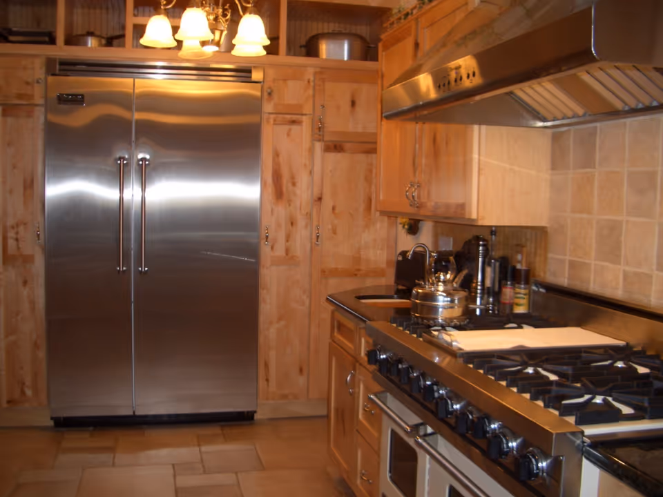 A kitchen with wooden cabinets, a large stainless steel refrigerator, a gas stove with multiple burners, a stainless steel range hood, and various kitchen items on the counter including a kettle and spice containers. There is a chandelier with five lights hanging from the ceiling.